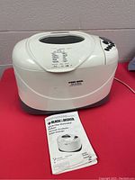 Photo of Black and Decker bread maker placed on red surface showing front and top with menu buttons and clear viewing window. Instruction manual placed beside the unit.