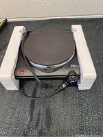 Photo of black Elite Cuisine countertop electric burner in original packaging with white foam side supports, showing cord and control knob.