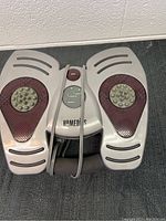 Top and front view of the silver and maroon Homedics foot massager showing the two foot areas with raised reflexology nodes and central controls including heat button and intensity dial labeled 'Homedics'.