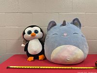 Both plush toys side by side on red surface in front of white wall with measuring tape showing size.