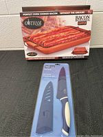 Gotham Steel Bacon Bonanza Copper pan box and InStyle chef knife in packaging on black surface against white wall.