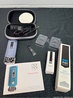 All items of the No! No! Pro Hair Removal system laid out on black cloth: hair removal device, power adapter, two treatment heads, carrying case, cut cleaner box, after treatment gel box, and instruction manual.