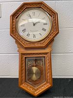 Full front image of the grandmother wall clock showing wooden case, clock face, and pendulum window.