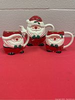 Santa Claus ceramic tea pot and two matching mugs shown on red surface against a neutral wall.