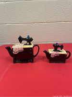 Photo showing two black ceramic sewing machine shaped teapots with detailed tablecloth and cloth draped