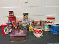 Collection of 18 assorted vintage tins in various colors and sizes arranged on floor, showing labels including honey, butter, and cigarette advertising.