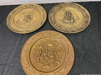 Three brass wall hanging plates displayed on a black fabric surface, showing two with ships and one with tavern figures.