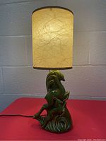 Full view of the vintage ceramic gazelle table lamp turned on, showing the lamp base and lighted cream fabric shade on a red surface against a white wall.