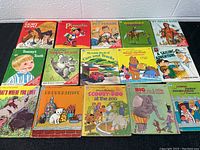 Photo shows 12 vintage children's books arranged on a black table, titles visible include The Goofy Goat, Pinocchio, Pet Parade, My Indian Book, and others.