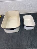 Two vintage rectangular metal medical trays, one larger and one smaller, placed on a dark carpet floor by a white wall.