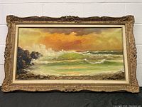 Frontal view of the large oil on canvas seascape painting, showing ocean waves crashing near rocks under a golden sky in an ornate gold frame.