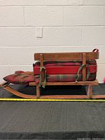 Side view showing wooden sled with plaid padded cushion and red rails, approximately 3 feet long with a measuring tape for scale.