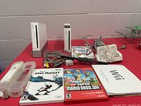 Wii remote with clear silicone cover shown alongside Epic Mickey and New Super Mario Bros Wii games, with Wii charging dock and cables behind.