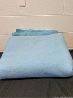 Two folded light blue wool blankets with stitched borders, stacked on black surface against white textured wall.
