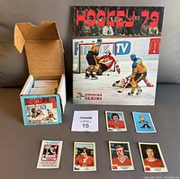 Photo showing the open box with multiple sticker packets inside, the 1979 Panini Hockey album and several loose stickers laid out including notable players and a cartoon character.