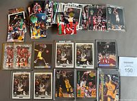 Overview of a large stack of basketball cards laid out on a surface with several cards in protective sleeves, featuring players in mid-action, some cards labeled as rookie cards.