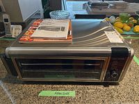 Front view of stainless steel Ninja Foodi digital air fry oven with clear door, control panel, and knob on the right side, sitting on kitchen counter with instruction manuals placed on top.