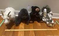 Four stuffed animal toys placed on wooden floor: white goat, black dog, black bear, clown doll.