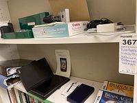 Shelf with Canon PowerShot SD1000 camera, box, manuals, Nintendo DS with manuals and software, Nook Color reader with case and manual, Sony speaker, batteries and various cables.