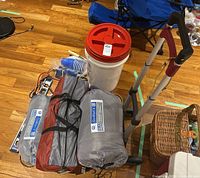 Overall view of camping gear including tents/sleeping bags, hand cart, storage container, plastic cups, gloves, and other camping accessories on wooden floor.