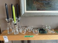 Photo showing three glass candle holders with tall black and green taper candles, two round glass candle holders, two green rectangular glass items, and two small glass figurines (horse and another animal) arranged on a wooden shelf.
