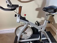 Side view showing the silver bike frame, flywheel, pedals, and black adjustable handlebar