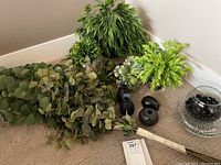 Photo showing multiple faux green plants including eucalyptus and ferns, four dark ceramic vases, and a clear glass vase with black and clear decorative stones showing the lot contents