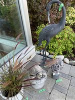 Shot showing the metal crane sculpture, resin owl figure, concrete turtle fountain, and ceramic pot with ornamental grass. Outdoor patio setting with stone pavers and greenery in background.