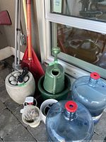 Photo of garden tools including red leaf rake and wooden spade, green ceramic watering can, concrete planter, white ceramic planters and two large water bottles.