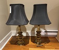 Two matching brass lamps side by side on wooden floor against a beige wall, with black shades and visible power cords.