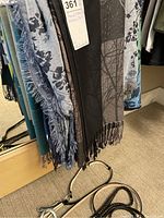 Various shawls and scarves with fringes and patterns hanging from a display, belts on the floor beneath