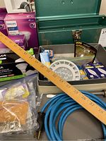 First photo showing wooden yardstick, blue extension cord coiled, assorted LED lighting products in packaging, flathead screwdriver, and hanging hardware near and inside a green metal toolbox.