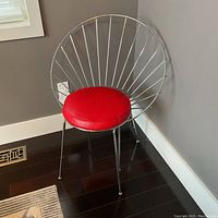 Front angle view of a 1950s chrome fanback chair with a round red vinyl seat cushion showing the distinctive sunburst metal back design and slender legs on dark wood flooring.