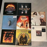 Top-down view of all 8 vinyl albums arranged in a 3x3 grid with one space empty; visible are Black Sabbath The Ultimate Collection box set, two KISS albums including Best of and Rocks Vegas, two Santana albums including Marathon and Africa Speaks, Paul Simon Graceland, and Lennon/Ono Greatest Hits.