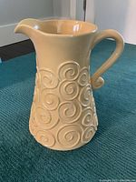 Front and side view of yellow ceramic pitcher with raised swirl design and curved handle on blue textile surface.