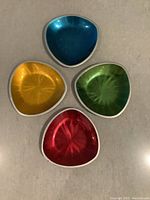 Four anodized metal dishes showing the colored etched interiors in blue, yellow, green, and red with triangular rounded shapes.