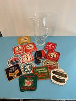 Full view of the clear glass cocktail pitcher alongside 16 assorted vintage bar coasters arranged on a blue surface.