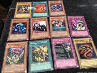 Photo showing 12 Yu-Gi-Oh! cards laid out on black fabric, including visible cards like Magical Marionette, My Body as a Shield, Worm Drake, and Castle Walls.