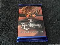 Front view of unopened 15-card caravan pack showing the title 'The Awakening' and series 'Legend of the Burning Sand' with distinct artwork and sealed blue edges.