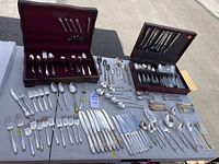 Wide view of both flatware sets laid out on table showing forks, knives, spoons, additional pieces, and two storage boxes.