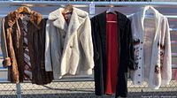 Four outerwear pieces hanging on metal rack: brown leather fur coat, off-white fleece coat, black velvet coat, and multi-color cardigan.
