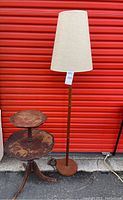 Full view of the wooden floor lamp with beige fabric lampshade standing next to the two-tier wooden accent table against a red background.