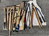 Wide view of the garden tools laid out on the ground, showing picks, axes, sledge hammer, shovel, and loppers.