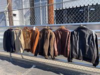 Wide view of all jackets and vest hanging on outdoor fence, showing different colors and materials including leather, suede, and corduroy.