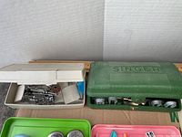 Singer buttonholer in green case next to white container with assorted sewing attachments, on cardboard background with measuring tape and half-yardstick behind.
