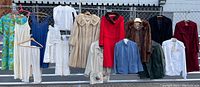 Overall view of clothing items hanging on fence including coats, jackets, dresses, nightgowns, housecoat, and children's clothes.