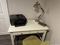 Photo of white desk with HP printer, adjustable desk lamp, three hole punch, and olive green chair underneath.