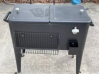 Side view of black drink cooler showing dual lids, bottle opener, wire rack,and tapered legs with wheels