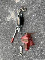 Small bench vise and 1000lbs load puller laying on asphalt surface; vise is red with screw handle; load puller has hooks and ratchet lever with red grip