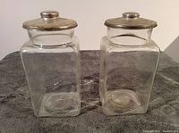 Photo showing two clear glass jars with aluminum lids, both jars upright on carpeted surface.